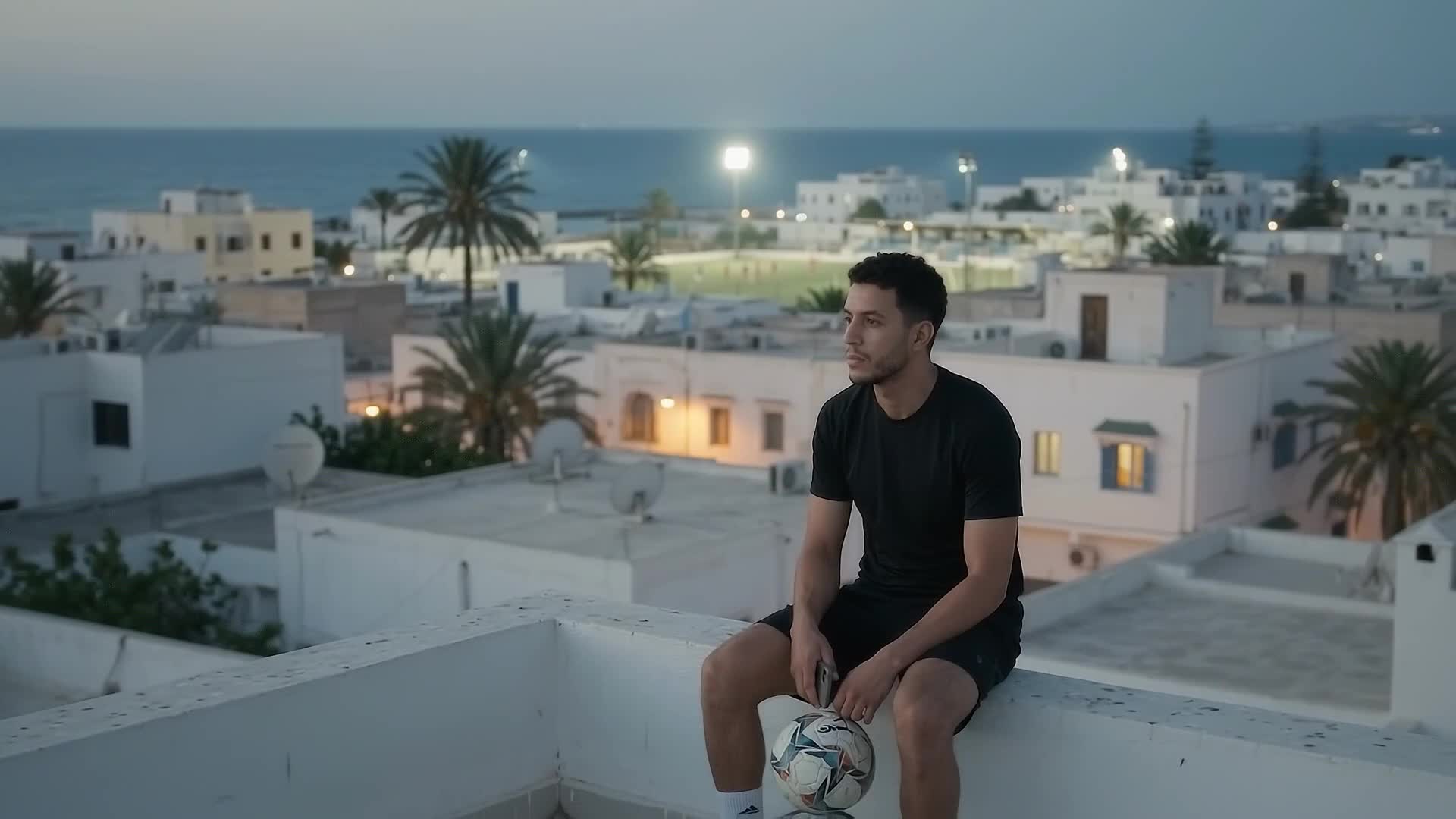 A player sitting on a Zarzis rooftop at dusk, ball at his feet and phone in hand