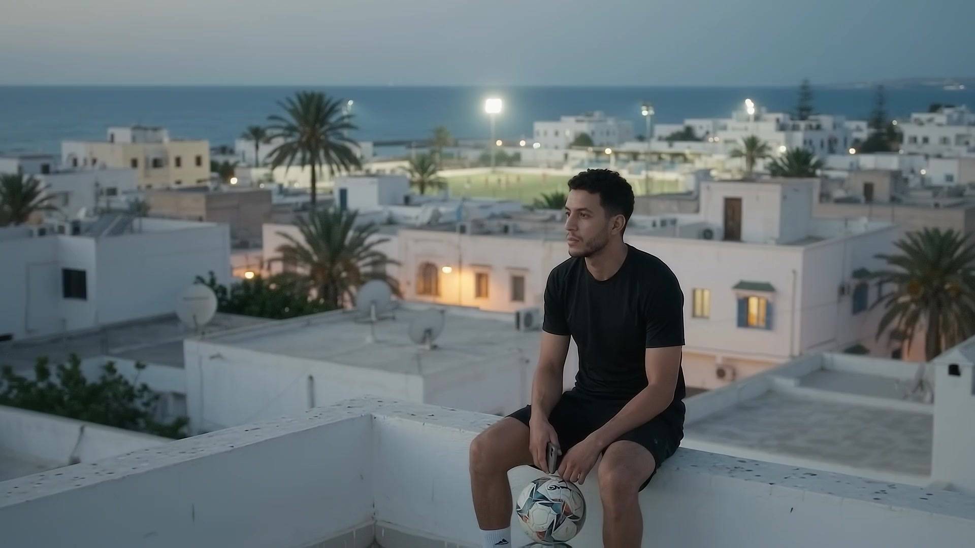 Un joueur assis sur un toit de Zarzis au crépuscule, ballon au pied et téléphone en main